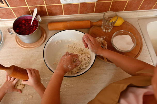Draufsicht auf die Küchenarbeitsplatte mit einem Topf karamellisierter Kirschen zum Füllen der Torten, Hände von Mutter und Kind kneten und rollen den Teig mit einem hölzernen Nudelholz aus. Selektiver Fokus - Foto, Bild
