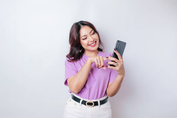 A portrait of a happy Asian woman is smiling and holding her smartphone wearing a lilac purple t-shirt isolated by a white background - Foto, afbeelding