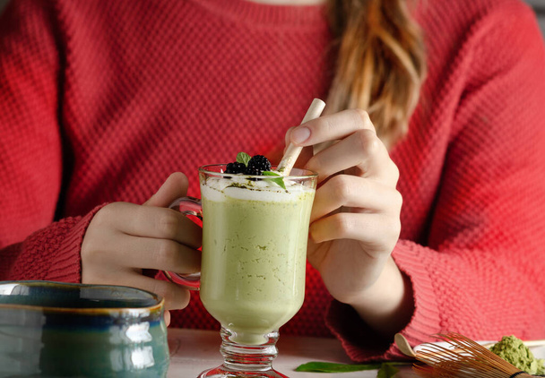 Weibliche Hand hält ein Glas mit einem Matcha-grünen Latte mit Schlagsahne und Blaubeeren. - Foto, Bild
