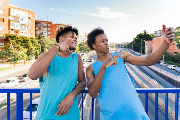 Two young afro american men wearing blue sleeveless t-shirt taking a selfie on the footbridge with the highway below in the city. - Photo, image
