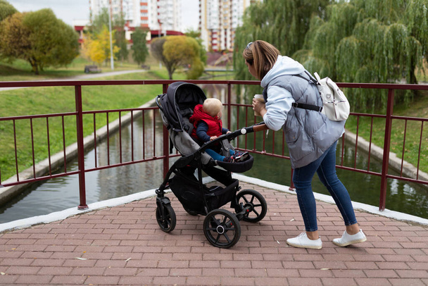 uma jovem mãe com uma mochila nos ombros caminha com seu filho em um carrinho de bebê no parque da cidade. - Foto, Imagem