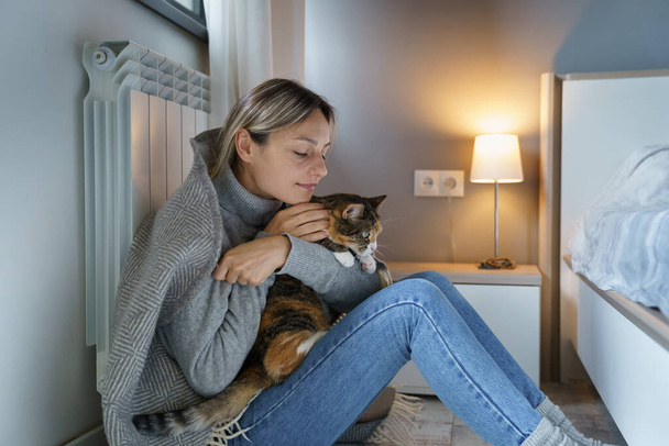 Young woman covered with warm plaid hugs favourite cat. Tired female owner enjoys playing with animal trying to be motivate after losing job. Cat makes blonde lady happy sitting near heating radiator - Фото, зображення