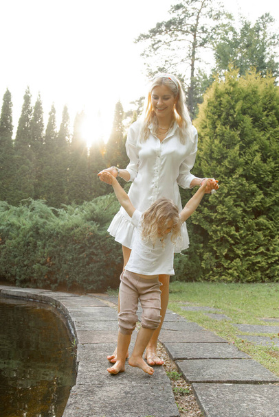 Vertical portrait of blonde barefoot woman and girl having fun, holding hands in nature outdoors. Nanny or sister, mother in family concept. Playing with offsprings, spend time on summer vacation  - Foto, afbeelding