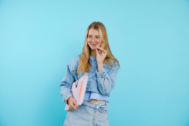 Confident, thoughtful, smiling blonde teenage girl in eyeglasses holding paper folder and biting pen in hands. Back to school, university. Education training course and class for students. Copy space - Fotografie, Obrázek