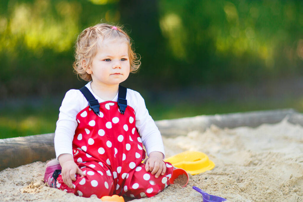Cute toddler girl playing in sand on outdoor playground. Beautiful baby in red gum trousers having fun on sunny warm summer day. Child with colorful sand toys. Healthy active baby outdoors plays games - Photo, Image
