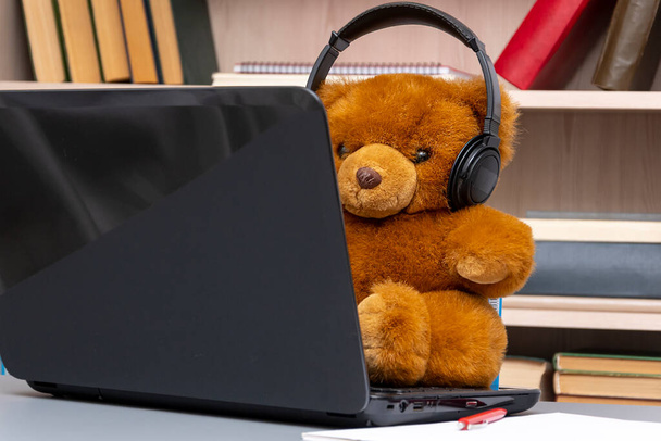 Toy teddy bear wearing headphones sitting at his laptop against a background of shelves of books and documents, close-up, selective focus. Concept: remote work, communication via the Internet. - Photo, Image