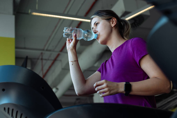 Tired overweight girl run on treadmill at fitness gym and quenching thirst drink water from bottle, from below view. Sweating woman do cardio workout on exercise machine. Weight loss, sport lifestyle. - Photo, Image