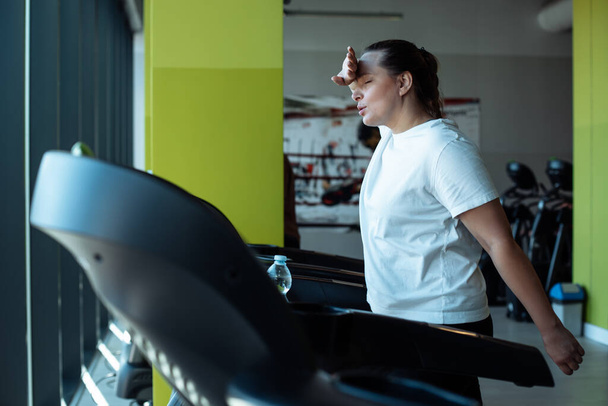 Tired plus size woman run on treadmill at fitness gym wiping sweat from face by hand with closed eyes, side view. 40s aged lady do cardio workout on exercise machine. Weight loss, sport lifestyle. - Photo, Image