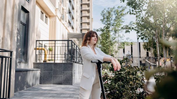 Beautiful young woman in white suit standing with her electric scooter near modern building in city and looking away - Фото, зображення