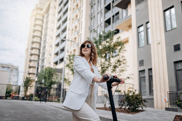Beautiful young woman in sunglasses and white suit standing on her electric scooter near modern building and looking away - Photo, image