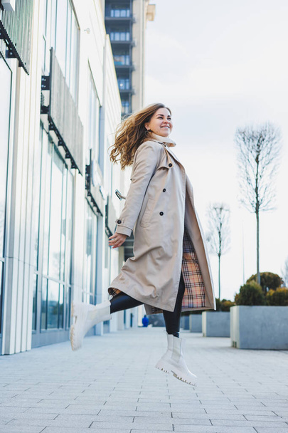Μια χαριτωμένη νεαρή γυναίκα με ένα μπεζ αδιάβροχο και φθινοπωρινές δερμάτινες μπότες περπατάει στο δρόμο. Φθινοπωρινή ζεστή εικόνα. Γυναικεία μόδα - Φωτογραφία, εικόνα
