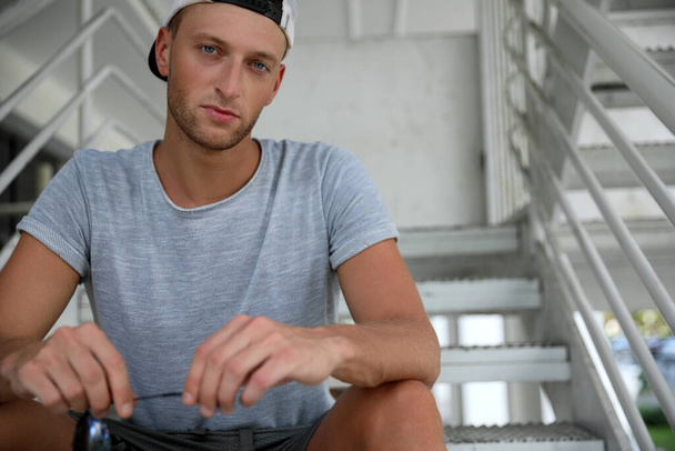 Handsome young man in stylish cap sitting on stairs outdoors. Space for text - Zdjęcie, obraz