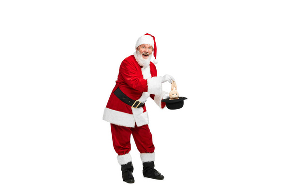 Portrait of senior man in image of Santa Claus showing tricks with rabbit and hat isolated over white background. Concept of fictional character, holiday, New Year, Christmas. Copy space for ad - Photo, Image