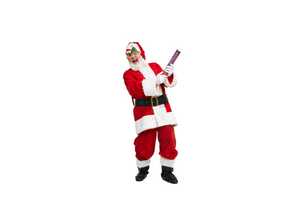Portrait of senior man in image of Santa Claus posing in festive glasses and confetti flapper over white background. Concept of fictional character, holiday, New Year, Christmas. Copy space for ad - Photo, Image