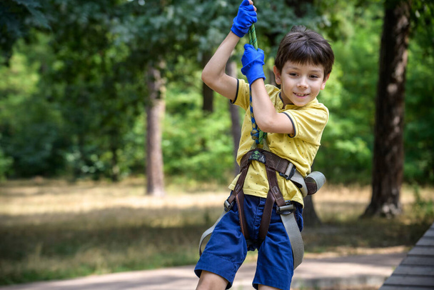 Strong excited young boy playing outdoors in rope park. Caucasian child dressed in casual clothes and sneakers at warm sunny day. Active leisure time with children concept. - Photo, Image