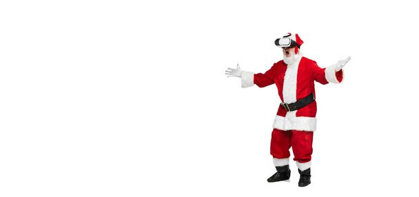 Portrait of senior man in image of Santa Claus posing in VR glasses isolated over white background. Gift idea. Concept of fictional character, holiday, New Year, Christmas. Copy space for ad - Fotografie, Obrázek