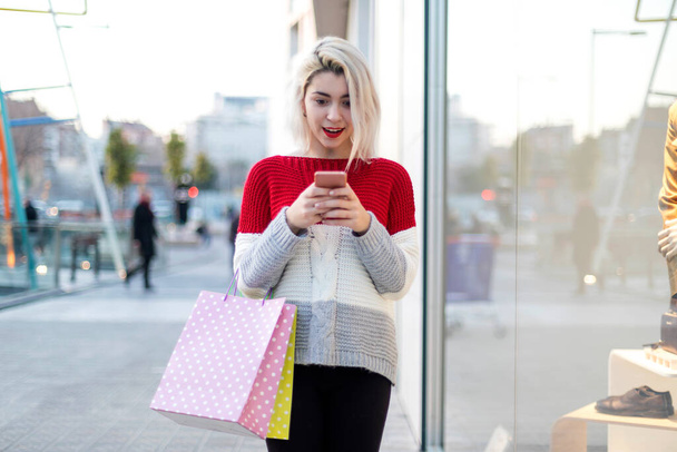 Front view of a young woman standing in a shopping center while using a mobile phone - Фото, изображение