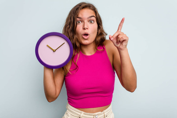 Young caucasian woman holding a clock isolated on blue background having an idea, inspiration concept. - Foto, Bild