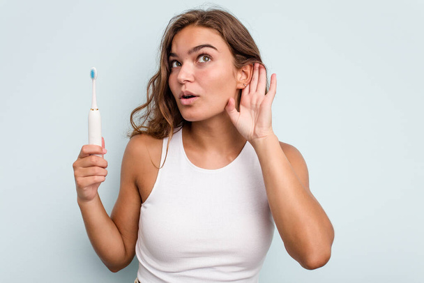 Jeune femme caucasienne tenant une brosse à dents électrique isolé fond bleu en essayant d'écouter un potins. - Photo, image