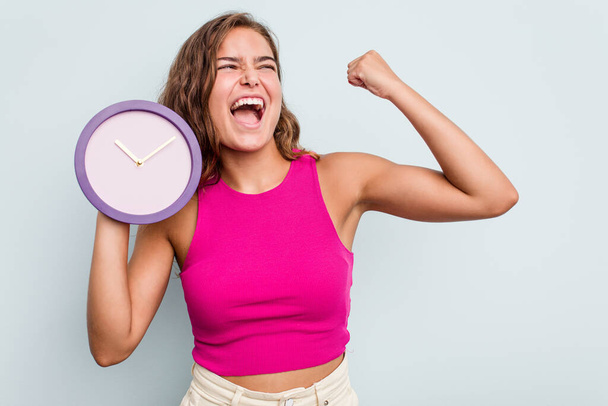 Young caucasian woman holding a clock isolated on blue background raising fist after a victory, winner concept. - Photo, Image