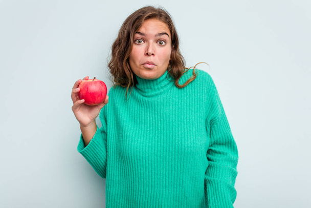 Junge kaukasische Frau mit einem Apfel auf blauem Hintergrund zuckt mit den Schultern und öffnet verwirrt die Augen. - Foto, Bild