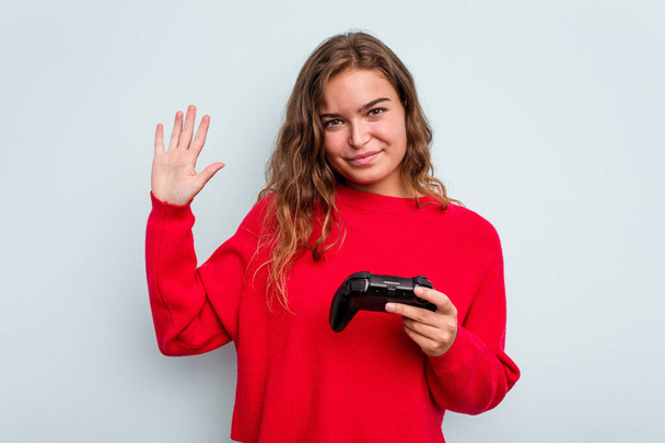 Junge kaukasische Spielerin mit einem Spielcontroller auf blauem Hintergrund, der fröhlich lächelnd die Nummer fünf mit den Fingern zeigt. - Foto, Bild