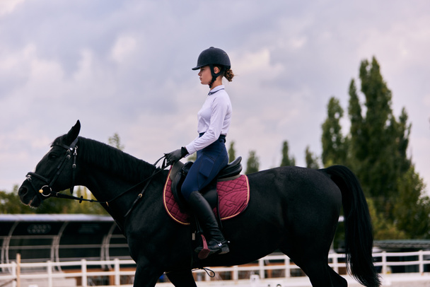 Equestrian sport. Portrait of teen girl, beginner female rider training at riding arena in autumn day, outdoors. Dressage of horses. Horseback riding. Concept of sport, grace, ad, active lifestyle - 写真・画像