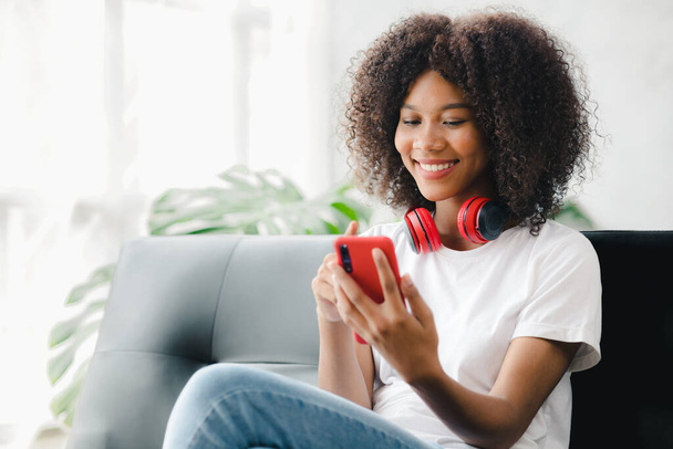 An American woman sits on the sofa on her smartphone, spends her weekends on social media, watching movies and listening to music at home. Holiday concept. - Foto, Bild