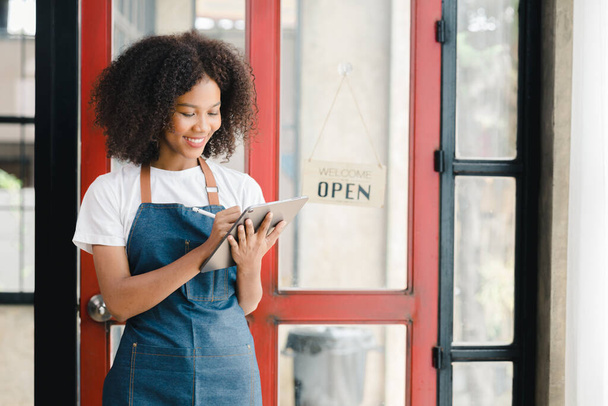 若いアメリカ人女性がオープンサインを持つレストランのドアの前に立って、彼女は顧客にサービスを提供するために店を開く準備をしているファーストフードレストランのウェイトレスです。レストランコンセプト. - 写真・画像