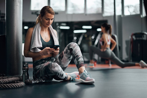 Pretty woman working out in a gym, making pause and using smartphone. Adult pretty sporty lady with beautiful shaped body. - Fotografie, Obrázek