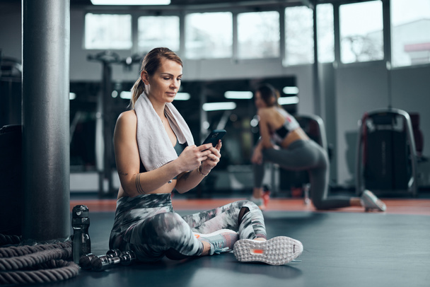 Pretty woman working out in a gym, making pause and using smartphone. Adult pretty sporty lady with beautiful shaped body. - Φωτογραφία, εικόνα
