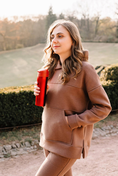 Молода жінка в светрі посміхається і тримає червоний термо-кухоль в осінньому парку. Сонячна погода. Осінній сезон
. - Фото, зображення