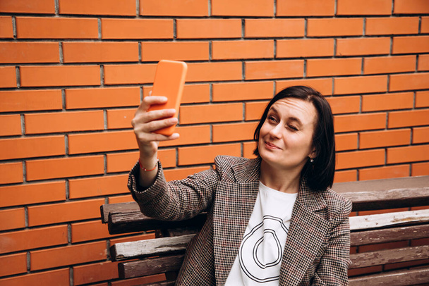 Une fille vêtue d'une veste brune s'assoit sur un banc et prend un selfie avec un œil fermé. Portrait d'une fille aux cheveux bruns sur fond de mur de briques rouges - Photo, image