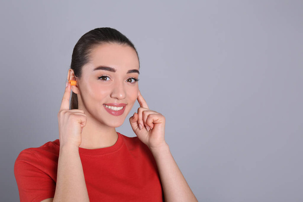 Young woman inserting foam ear plugs on grey background. Space for text - Photo, Image