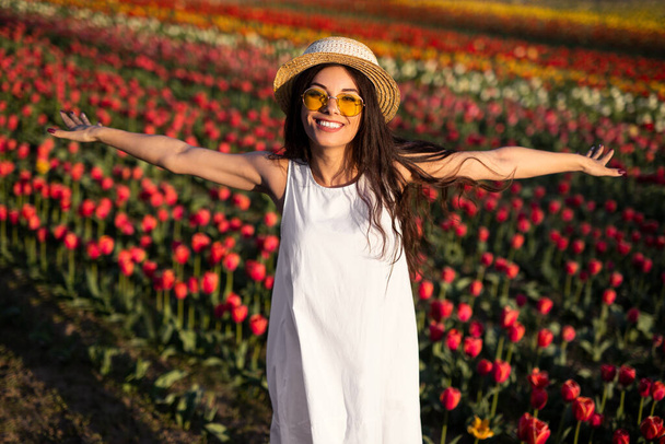 Delighted young female tourist in white dress straw hat and sunglasses, smiling and looking at camera while standing in tulip field during trip in Netherlands - Valokuva, kuva