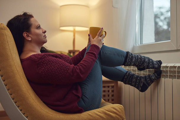 Frau heizt Füße an einem kalten Wintertag, hält Kaffeetasse, Energie- und Gaskrise, Kühlraum, Heizungsprobleme. - Foto, Bild