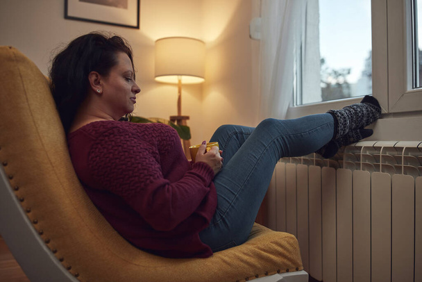 Женщина нагревает ноги в холодный зимний день, держа чашку чая кофе, энергетический и газовый кризис, холодная комната, проблемы с отоплением. - Фото, изображение