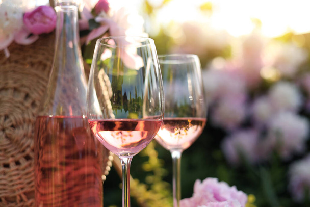 Bottle and glasses of rose wine near straw bag with beautiful peonies in garden, closeup - Foto, Imagem