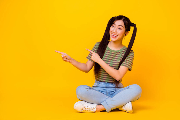 Photo of young attractive woman sitting on floor point fingers blank space show advertisement isolated on yellow color background. - 写真・画像
