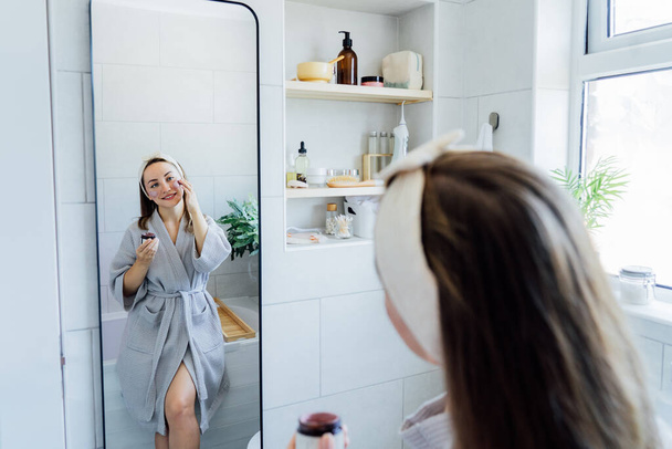 Young woman in bathrobe looking in the mirror and applying facial natural cosmetic clay mask on her face in bathroom. Cosmetic procedures for skin care at home. Beauty self-care. Selective focus - Zdjęcie, obraz