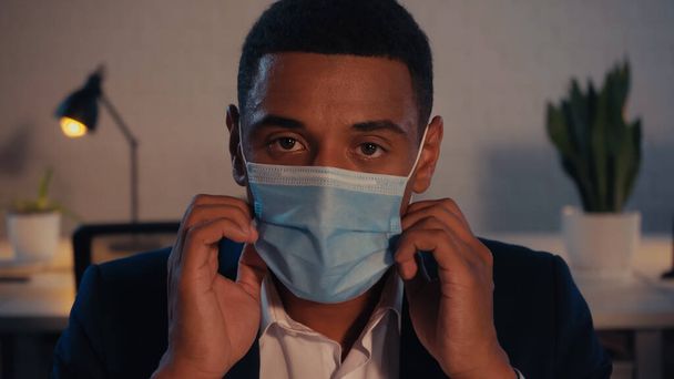 Afro-Amerikaanse zakenman in medisch masker op zoek naar camera in het kantoor 's nachts - Foto, afbeelding