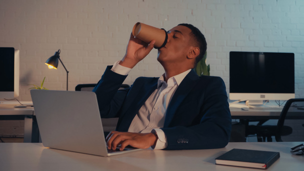 Afrikai amerikai menedzser öltönyben kávét iszik és laptopot használ éjszaka az irodában. - Fotó, kép