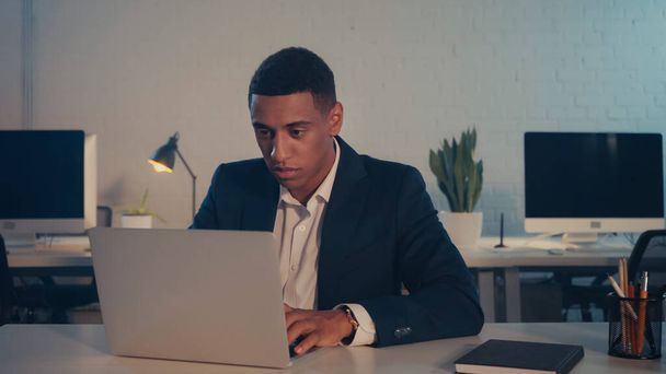 Afroamerikanischer Geschäftsmann nutzt Laptop in der Nähe von Notebook in der Nacht im Büro  - Foto, Bild