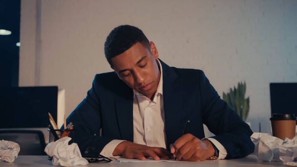 African american businessman writing on paper near coffee to go in office at night - Foto, Imagen