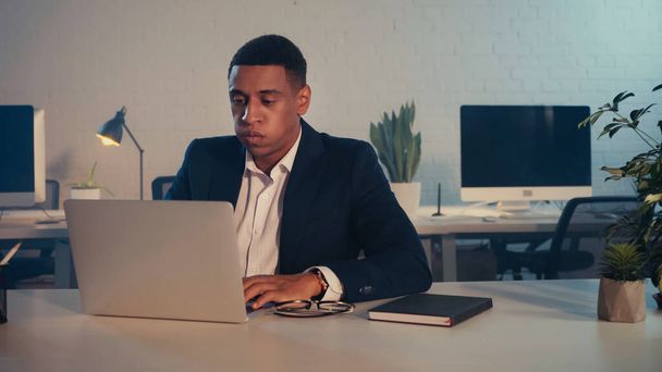 Exhausted african american manager using laptop near eyeglasses and notebook in office at night - Photo, image