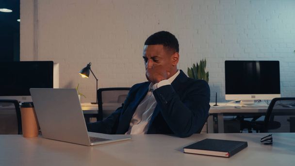 Exhausted african american businessman in suit sitting near laptop and takeaway drink in office  - 写真・画像