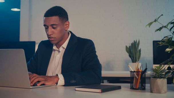 Uomo d'affari afroamericano che utilizza laptop vicino notebook e piante in ufficio di notte  - Foto, immagini
