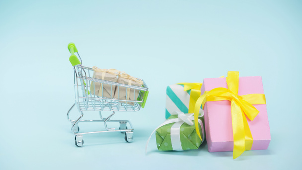 small shopping cart near wrapped presents on blue background - Фото, изображение
