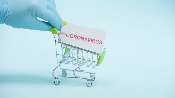 vista cortada de pessoa em luva de látex segurando carrinho de compras com letras coronavírus no cartão no fundo azul - Foto, Imagem