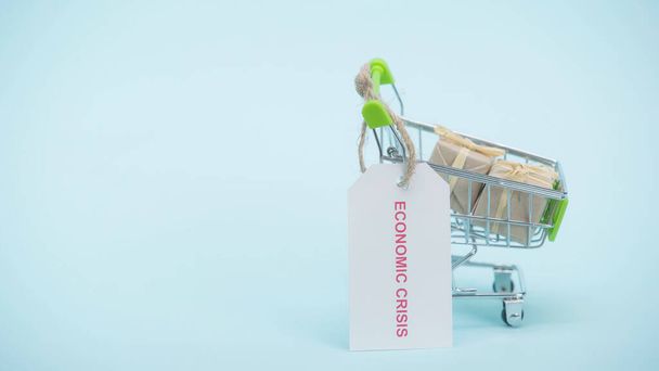pequeño carrito de compras con regalos y tarjeta con letras de crisis económica sobre fondo azul  - Foto, imagen